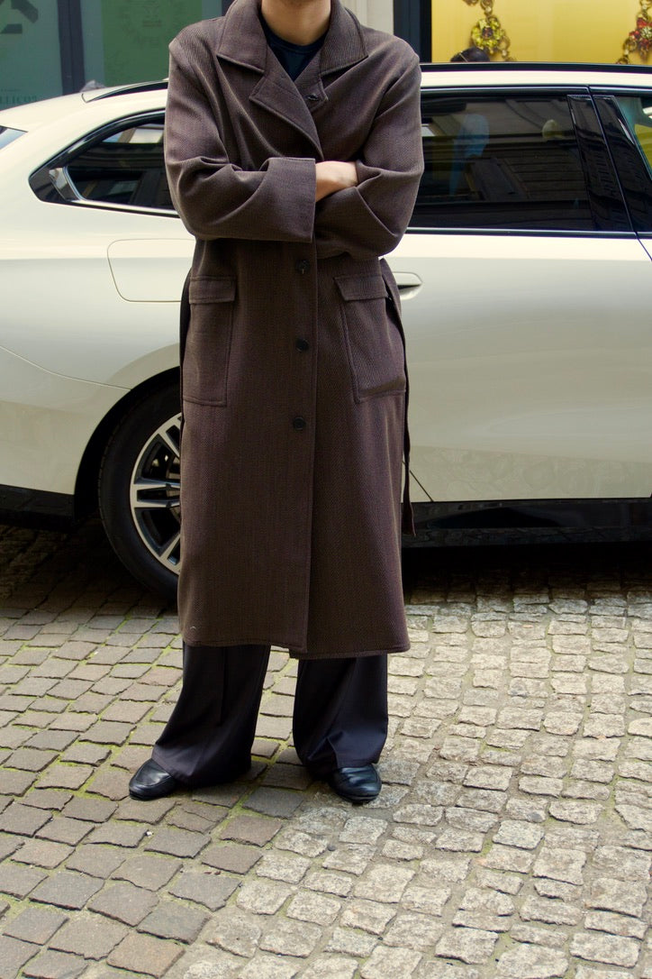 A person wearing a long brown coat, black pants, and black shoes stands with arms crossed in front of a white car on a cobblestone street.